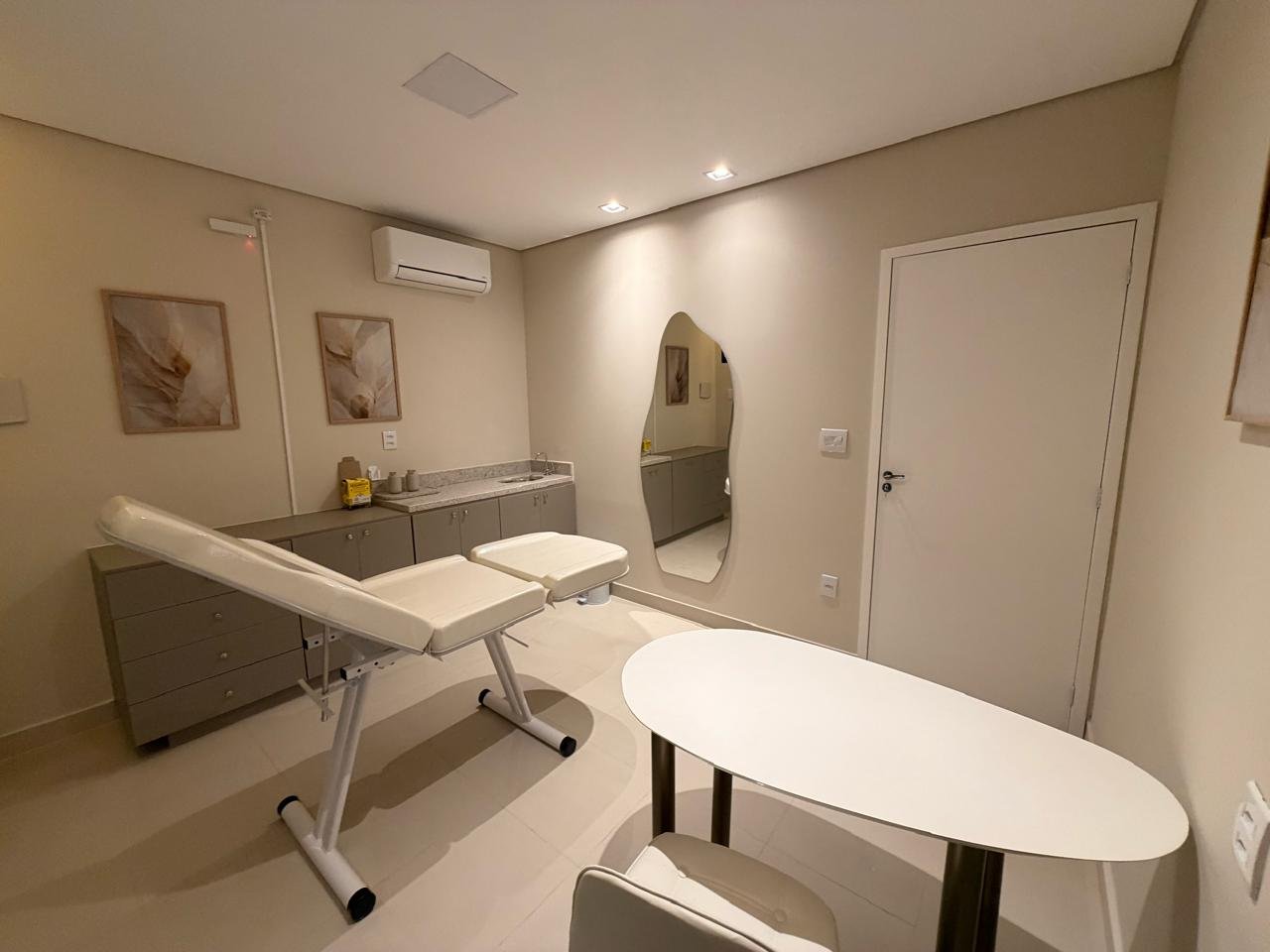 Sala 03: consultório com maca branca, bancada com pia, espelho de formato orgânico, mesa oval e ar-condicionado